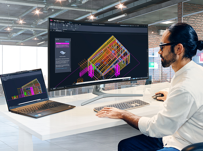 Person working on a computer and laptop both displaying a 3D house model in CAD software.